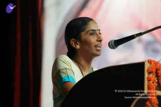A woman speaks into a microphone at a black lectern decorated with orange marigold garlands. She wears a cream sari with a turquoise and gold-embroidered border, a black beaded mala necklace, a small red bindi, and stud earrings, her hair pulled back. Behind her a large backdrop shows a blurred portrait of a figure wearing an orange-pink turban. Dark red curtains frame the left side of the stage.