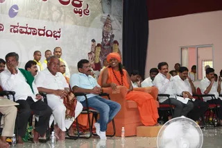 A seated saffron-robed figure with an orange turban and long dark hair, wearing a rudraksha mala, sits smiling in an orange armchair on a stage. Probable Swamiji by appearance cues: orange turban, saffron robes and rudraksha mala on the central throne-like seat. Around him sit men in white shirts and dhotis, a man in a light blue shirt and jeans, and men in dark trousers and striped shirts, some with a green shawl and others with a gold-bordered wrap. Behind them stand shaven-headed men in white with yellow shawls. Behind the stage, a large banner shows a deity murti with garlands and Kannada script. A white fan and water bottles sit in the foreground.