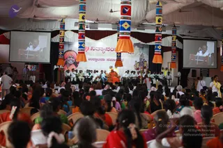 A large indoor hall with a white draped ceiling hosts a seated audience of many women in colourful saris, mostly from behind, with some in red, orange and green. At the front, a central figure in an orange robe and orange turban sits on a raised stage with a group of people dressed in white seated along both sides. Two large screens on either side show a speaker at a white podium. Tall decorated pillars with bold geometric patterns and orange tassels frame the stage. A backdrop banner shows a smiling figure in an orange turban, a deity image and a line of script. a person: the central figure wears an orange robe and orange turban, a saffron-toned appearance consistent with the page's claim, though the face is small and distant.