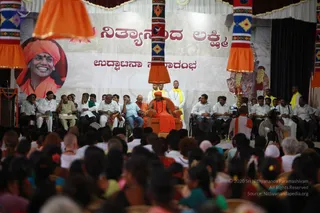 A large public satsang on a stage draped in maroon and gold, with a backdrop reading Nithyananda Lakshmi and a smiling portrait of a man in orange turban at left. A central figure in orange robes and a rudraksha mala sits in a large orange-upholstered chair, flanked by seated men in white shirts and dhotis, and attendants in white and yellow shawls standing behind. A silver lamp stands on a stand near the chair. Ornate orange-and-patterned banners hang from the top corners. In the foreground, a dense audience of several hundred people, many women in colourful saris, sits facing the stage. A deity mural is visible at right.