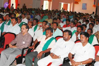 A large seated audience in a hall with orange-peach walls and red-cushioned chairs, all facing forward toward an unseen speaker. In the foreground, several men sit close together: a man in a checked plaid shirt and grey trousers with an ID badge on the left, a man in a white shirt with a green shawl, a man in a white dhoti-style outfit and watch, and a man in white with a green shawl on the right. Many other attendees in white, pastel and bright shirts fill the rows behind, some with green shawls, and a few people stand near the back wall. A blue wall sign is visible at upper right.