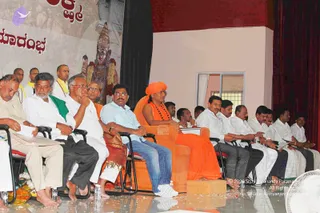 A stage event with seated dignitaries in a row of chairs. In the centre, a person wearing a saffron-orange turban, saffron-orange robe and a rudraksha-style necklace sits on an orange throne-like seat, holding a small white paper. Flanking figures wear white dhotis and shirts, one in a light blue shirt, another with a green shawl. A framed deity murti with garlands and a red-and-gold Kannada-language banner stand behind the stage on the left. Several younger men in white and yellow robes stand at the back. Water bottles and fruit offerings sit on the floor, and a fan is at the lower right. Swamiji (probable) is seated centre on the orange throne, wearing a saffron turban and saffron robe with a rudraksha necklace.