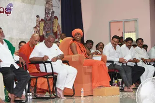 A photograph of a seated assembly at an indoor satsang hall. In the centre, a person in an orange saffron robe and an orange turban sits in a tall orange-upholstered armchair with a rudraksha mala around the neck, smiling and looking to the side. Beside them, a bald man in an orange robe sits on a red chair, and a man in a white shirt and white dhoti writes on a sheet. Other men in white shirts and dark trousers sit in rows of chairs. A white ceiling fan and water bottles are at the front, and a painted deity murti with flower garlands stands on the backdrop. The floor is strewn with petals.