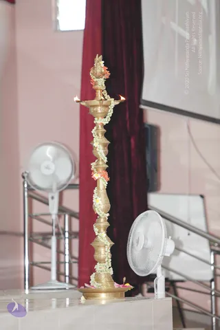 A photograph of a tall polished brass standing oil lamp (nanda vilakku style) on a white stage platform. Its multi-tiered stem is wrapped with white jasmine-type garlands and small orange and white flowers, and flame-lit wicks sit on the lamp's wide bowls with a bird-shaped finial at the top. Pink and white petals are scattered on the base. Behind it hang a deep maroon velvet curtain and a pale pink wall, a projector screen is at upper right, and two white pedestal fans stand on the stage. No people are visible.