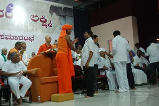 A photograph of a stage event with a large backdrop showing Kannada script and the word Lakshmi in red. A figure in a saffron-orange garment with a saffron head cloth stands centre-stage, reaching out to shake hands with a man in a white shirt and dark trousers. Beside an orange-upholstered throne-style chair, a shaven-headed man in orange robes holds a garland. Men in white dhotis and shirts sit on red-cushioned chairs and stand around the stage, with flower petals scattered on the white floor.