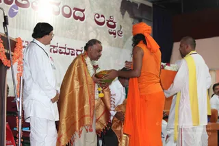 A stage event with a banner behind reading Nithyananda Lakshmi in Kannada script. A man in a white kurta with a gold-bordered shawl and white flower garland bows forward, receiving a brass plate or bowl of offerings from a figure in a saffron-orange top and matching orange head wrap with long dark hair. A man in white with a yellow shawl stands at right holding a bouquet of white flowers. A standing man in white with eyeglasses and a lanyard stands at left beside an orange-draped wooden podium with marigold garlands. Other seated attendees are partially visible in the background.