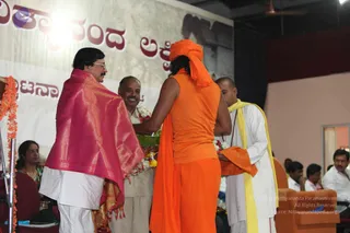 A stage event photographed from behind a figure in a saffron-orange sleeveless top, saffron dhoti and orange turban with long dark hair falling below it. This person holds a green tray of offerings and flowers toward a man in a pink and gold silk shawl over white clothes, who stands at left with a moustache and glasses. A third man stands in the centre in beige clothes wearing a flower garland. At right, a bald man in white with a yellow shawl and rudraksha mala stands beside an orange cloth, and seated audience members in white shirts sit in orange armchairs. Behind them, a backdrop shows large red Kannada lettering and a stylised painted figure. Marigold garlands hang at the left edge.