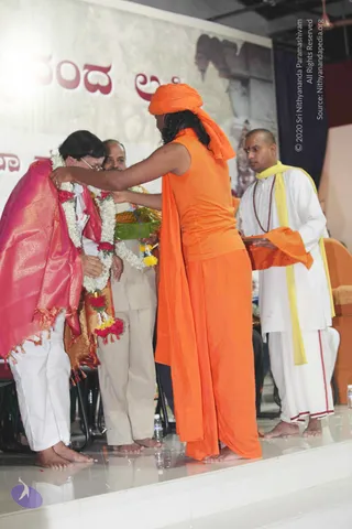 Photograph on a stage with a glossy white floor. A figure in an orange turban, sleeveless orange top and orange dhoti with long dark hair and a flowing orange shawl stands with back to camera, placing a garland of white and red-yellow flowers on a man in white shirt and white trousers who wears a pink-gold shawl and glasses and bows his head. A second man in beige shirt and trousers stands behind. On the right a shaved-headed man in white kurta, white dhoti and yellow shawl with rudraksha mala holds an orange cloth and a tray. Behind the stage a banner shows Kannada script in red and a large cutout portrait. Water bottles sit on the floor. The orange-clad figure is a probable Swamiji by appearance cues (long swept hair, orange turban and clothing), so this is an in-person candidate.