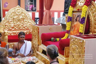 A photograph of a Paduka Puja ceremony. Ornate gold-carved thrones with red cushions stand on a golden platform. On the right, a framed portrait of a figure in saffron/orange robes with a long mala necklace is garlanded with yellow, orange and purple flowers, and sits on the red cushion of a throne. In the centre-left, a shaved-headed priest in a white dhoti with bare chest, white body markings and a rudraksha mala sits before the gold platform, which holds silver trays of flowers and offerings. Two onlookers in the foreground, one wearing a dark beaded necklace, watch from behind the trays. An orange and red background wall with a printed board is behind.