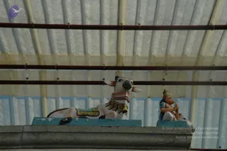 A photograph of two painted sculptures on a concrete ledge beneath a translucent corrugated roof with metal rods. On the left, a white recumbent bull with a pink and gold garland and golden decorated saddle cloth lies on a turquoise plinth. On the right, a small figure with a gold crown, turquoise garment and gold bangles sits with hands pressed together in prayer on a matching turquoise plinth. No physical people are visible.