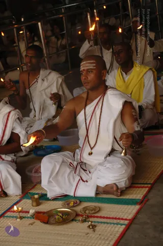 A photograph of a Hindu ritual in a hall with metal railings and several oil lamps held aloft by seated participants. In the centre, a shaven-headed priest in plain white dhoti and shawl with red borders, wearing a rudraksha mala and white sacred ash lines on his forehead, holds a lit flame in his right hand and a brass bell in his left. Before him on a woven mat are brass plates with flowers and fruit, a small brass cup, and a standing brass oil lamp. Behind him, other men in white garments, one in a yellow shawl, sit with lamps and bells. Crowds of seated devotees fill the background.