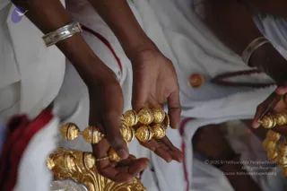 Close-up photograph of two pairs of hands reaching toward a set of ornate gold beaded items. The people wear white clothing with red-bordered cloth, and one wrist has a silver bangle. Hands and gold beads dominate the frame.