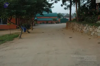 A photograph of a dirt road running through an ashram-style grounds with no people visible. On the left, a leafy tree stands beside an orange-painted building, a blue plastic barrel and small potted plants near its base. Ahead, an orange building with green corrugated roofs sits beyond a green metal gate, and a thatched-roof hut appears in the distance. On the right, a dry-stone retaining wall borders the road, with flowering shrubs and a tree behind it. A partially visible white sign hangs at the upper right edge.