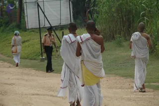 Outdoor photo on a dirt path beside grass and tall reeds. Three shaven-headed men in white dhotis with red-bordered cloth walk away from the camera; the middle one, in a yellow undergarment, holds a mobile phone to his ear and wears a rudraksha-style bead necklace. A uniformed security guard in khaki and black trousers walks in the background, and a woman in a white sari carrying a bag walks on the left. A large blank signboard on a metal frame stands behind them.