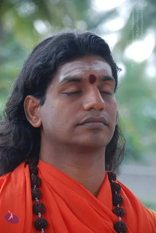 Close photo of a man with long dark hair, eyes closed, face lifted slightly in meditation. He has a white vertical tilak with a red kumkum dot on his forehead, wears an orange-saffron shawl draped over the shoulders and a multi-strand rudraksha mala. Blurred green foliage in the background. a person by appearance cues: long swept-back hair, saffron clothing and rudraksha malas, shown in meditation.