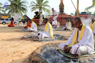 A photograph of a ritual fire ceremony outdoors on sandy ground beside a temple. Several men in white dhotis and yellow shawls sit cross-legged on mats and in front of fire pits filled with grey ash, their eyes closed in meditation or prayer. A monk in orange robes sits further back at left, and a man in white stands near a tall copper-coloured kalasha with a yellow-orange floral garland, with a black Shivalinga-style stone nearby. A silver SUV is parked at left, and palm trees and smoke rise in the background. A printed board stands on a pole at left.