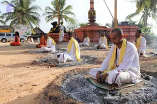 Outdoor photo of a row of men seated cross-legged on mats in front of small shallow fire pits filled with grey ash, on bare reddish ground beside coconut palms. In the foreground a man with a shaved head wears a white dhoti and white top with a yellow shawl and a beaded mala, eyes closed. Behind him, several seated men wear white with yellow shawls, and a few in orange robes sit further back. A standing man in white with a yellow shawl stands near a tall ornate copper-coloured kalasha pillar with a garland, beside a dark stone deity murti and a red stepped platform. A signboard and a parked silver SUV appear at the left.