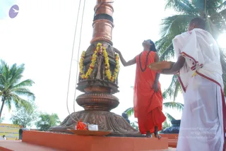 Outdoor photo of a copper flagstaff with a bronze base, set on an orange-tiled platform beneath palm trees and a bright sky. A garland of yellow and red marigolds and a bundle of straw hang on the pole. In the centre, a figure in a saffron-orange robe and a rudraksha mala stands barefoot, one hand raised to touch the flagpole and face tilted upward. To the right, a man in a white dhoti with red-bordered shawl holds a brass plate. Yellow banner text and a small orange cloth sit at the base.