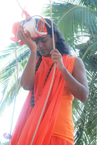 A man in an orange sleeveless top and orange wrap with a black rudraksha mala hangs on a rope, holding a bundled orange, white and blue flag in one hand while looking down. Palm fronds fill the background. The bundled flag is hoisted on the rope as part of a flag-hoisting ceremony.