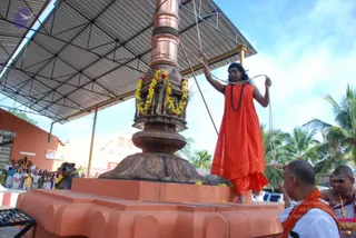A man in a saffron-orange robe stands on a raised orange platform beside a tall copper flagstaff wrapped with copper bands and garlanded with yellow and red marigold flowers. He holds a rope attached to the pole with both hands, as if hoisting a flag. A rudraksha mala hangs at his chest and his long dark hair falls past his shoulders. Below, a man in white with orange shawl stands by, and a priest with a shaved head and white attire holds a small brass vessel. A row of people in white and blue clothing stands under a corrugated metal roof on the left, beside a sign reading TEMPLE. Palm trees and blue sky fill the background.