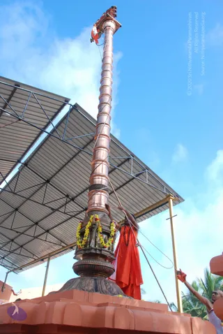 A tall copper flagstaff, wrapped in twisted copper bands, rises from an ornate stone base strung with yellow and orange marigold garlands, topped by a small carved finial and a saffron flag. A figure in an orange-red robe and a rudraksha mala hauls on a rope beside the pole, while another person in white reaches up from the lower right. A corrugated metal roof shelter and a blue sky with palm trees frame the scene.