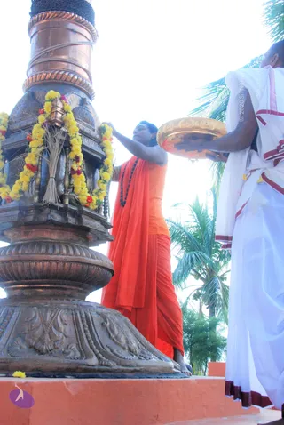 A photograph of a flag-hoisting setup beside a tall ornate copper-banded pole with a carved bronze base, garlanded with yellow marigolds and red flowers, with dried straw bundles tied at its foot. A man in a saffron-orange robe and dark rudraksha mala reaches up to adjust the garland on the pole. Beside him, a man in white dhoti and white shirt with red-bordered cloth holds a large brass plate. Palm trees and bright sky fill the background, and the scene is shot from a low angle on an orange-tiled platform.