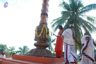 Outdoors under a bright sky beside tall coconut palms, a tall copper flagpole rises from an ornate bronze base wrapped with yellow and orange marigold garlands. A figure in a long orange-saffron robe with long, swept-back dark hair and a rudraksha mala stands on an orange stone platform, holding up a small object. Two men in white dhotis with maroon borders stand beside him, one holding the hand of the other, both facing the pole. Swamiji (probable): the saffron robe, long hair and rudraksha mala are visible cues, so the central figure is a probable candidate for Swamiji performing the flag hoisting ritual.