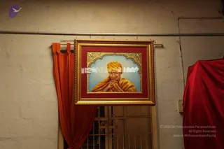 A framed painting hangs on a whitewashed wall above an orange curtain and a doorway with a grille. The gilt frame has a red mat inside and an ornate arched gold border around a painted portrait of a man in a saffron turban and saffron shawl, hands pressed together beneath his chin against a pale blue background. A red cloth drapes over an object at the right edge. The portrait depicts a person and is not a live individual; no physical people are present. A cropped ceiling bracket and pipe run across the top. Copyright text overlaps the lower right corner.