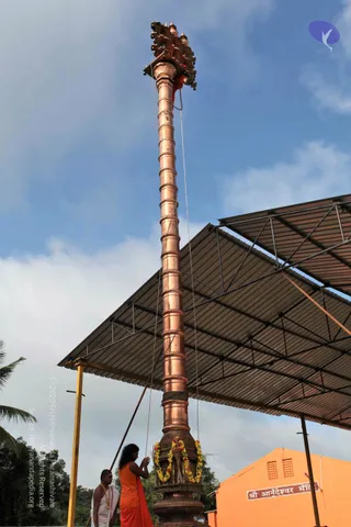 A tall copper flagstaff, ringed with bands and topped with an ornate ornamental finial and a red cloth, rises against a blue sky with clouds. Its base is wrapped with a garland of orange and yellow flowers. A long-haired person in an orange saffron robe stands at the base holding ropes that raise the pole, while a second man in white dhoti and shawl with a beaded necklace assists beside a corrugated metal canopy. Behind them an orange building bears a sign reading Sri Anandeshwar Mandir.