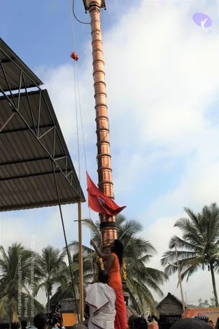 A tall copper flagpole rises against a pale blue sky with scattered clouds, with coconut palms along the edges. A red flag with a blue emblem is being raised on the pole. A woman in an orange sleeveless top and orange trousers stands on a raised platform, pulling a rope to hoist the flag, while a man in a white dhoti with a red border stands below her. A corrugated metal canopy stands at the left edge, and a small red bundle hangs from a rope near the top. Several spectators are visible at the bottom, and a small shrine structure and a poster with printed text appear at the lower right.