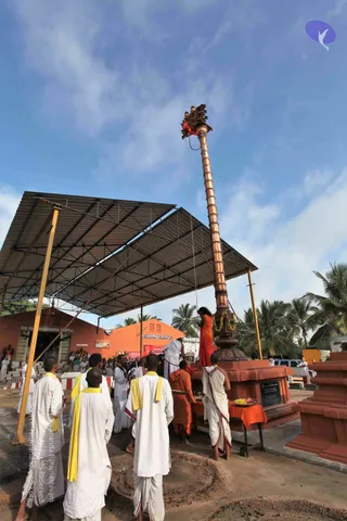 Outdoor flag-hoisting ceremony at a temple courtyard beneath a corrugated metal shelter. A tall copper flagstaff with a decorated finial rises against a blue sky, with a red flag attached near the top. A figure in orange robes stands at its base, holding the rope, while a man in white dhoti and shawl stands nearby. A group of men in white dhotis with yellow shawls stand in the foreground watching. Behind them, an orange building labelled Sri Anedeshwara Temple and a shrine with palm trees complete the scene. A table with orange cloth holds a plate of flowers and offerings. Several people, including figures in white, stand in the background.