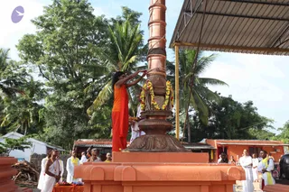 Outdoor photo of a flag-hoisting ceremony beside a tall copper flagstaff set on an orange-painted stone base. A figure in a saffron-orange top and wrap, with long dark hair swept back, reaches up with both arms to adjust a garland of yellow and orange marigolds at the top of the pole. Nearby, men in white dhotis and yellow-and-white shoulder cloths stand around the platform, some holding garlands and offering trays with flowers and fruit. Palm trees, a thatched shelter with a metal roof, and orange-walled buildings surround the scene.