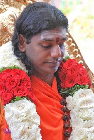 A man with long dark hair sits with eyes closed in a gilded throne-style chair, wearing an orange-saffron shawl over his shoulders and a dark rudraksha mala. He wears red and white rose garlands around his neck and a red tilak on his forehead. Behind him is ornate gold carving and soft blurred floral background. Swamiji (probable): long swept-back hair, saffron clothing, rudraksha mala and garlands are visible cues, with the event page indicating his presence.