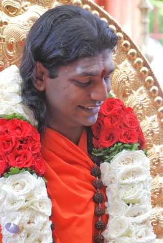 Close photo of a smiling man with long, swept-back dark hair, eyes lowered, wearing an orange-saffron garment and a rudraksha mala across his chest. He has a red bindi and white vertical markings on his forehead. He is garlanded with red roses, white roses and green leaves, seated before an ornate gold-embossed throne with raised circular studs.