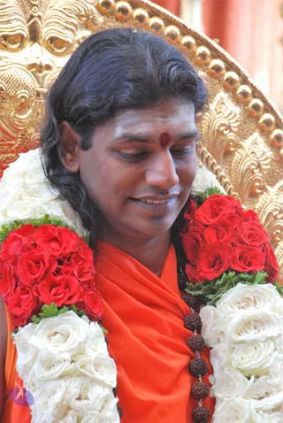 Close photo of a man with long swept-back dark hair and a red bindi on his forehead, smiling with eyes lowered. He wears an orange-saffron robe and a double garland of red roses and white roses with green leaves around his neck, with dark rudraksha beads on his chest. Behind him is an ornate gold throne with carved details and a red backdrop. Swamiji (probable): the long hair, saffron robe, rudraksha malas and gold throne are the cues for this figure seated in a place of honour.