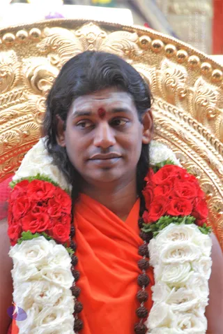 A man sits on an ornate gold throne with carved floral scrollwork and round gold finials. He wears long, swept-back dark hair, a red bindi and white tilak on his forehead, an orange-red saffron shawl, and a dark rudraksha mala. A thick garland of red and white roses with green leaves drapes over his shoulders.