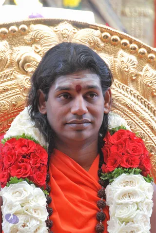 Close photo of a man seated before an ornate gold throne with carved floral scrollwork and round gold finials. He wears an orange-saffron shawl over his shoulders and a long rudraksha mala, with a red kumkum mark on his forehead. He has long dark hair swept back and is draped with a garland of red roses and white roses with green leaves. a person: the long swept hair, saffron wrap, rudraksha mala and ornate throne are the cues, and he is seated in person.