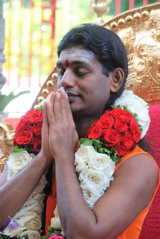 A photo of a man seated with eyes closed and palms pressed together in anjali mudra. He wears an orange sleeveless top, a red kumkum dot on his forehead, and a garland of red and white roses with green leaves around his neck. Behind him is an ornate gold throne with a red cushion. The setting appears to be an outdoor or semi-open space with blurred greenery and red structures. Identity uncertain from appearance alone.