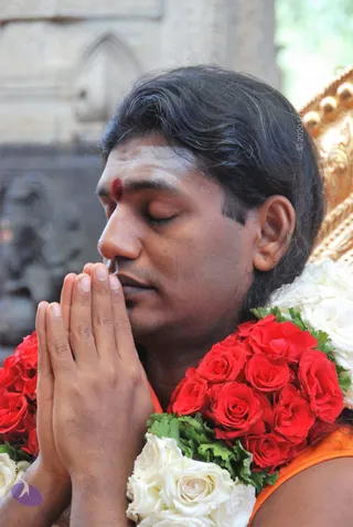 Close photo of a man with eyes closed and palms pressed together in anjali mudra at his chest. He wears a red bindi-like tilak on his forehead and an orange garment at his shoulders, with a large garland of red roses and white roses draped across his shoulders. Behind him is a gold-toned carved object and a stone temple wall.