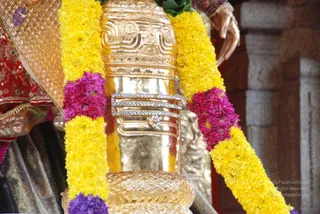 Close-up photograph of a gilded deity murti, with a polished gold cylindrical form decorated with raised scrollwork and a band of studded gems. Yellow marigold garlands and magenta flower strands drape across it. Behind it, a figure in red and gold embroidered garments with a gold shoulder ornament is partly visible, and a stone pillar stands in the background. No people are clearly visible in the foreground.