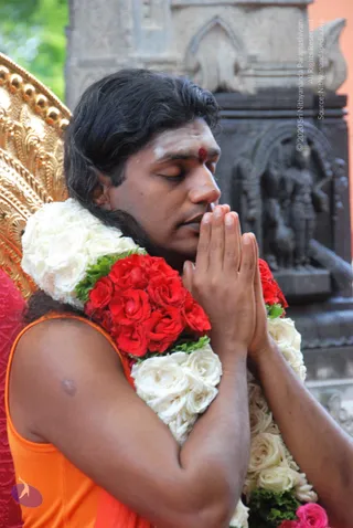 A man with shoulder-length dark hair, eyes closed, palms pressed together in namaskar at his chest. He wears a red bindi and white tilak on his forehead and an orange sleeveless top. A garland of red and white roses hangs around his neck and shoulders. Behind him is a gold-embossed ornate chair and a dark carved stone shrine panel with figures. The photo is a close portrait and shows one visible person.