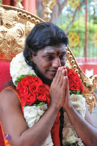 A photograph of a man with long dark hair seated in front of an ornate gold-colored throne with carved scrollwork and a red cushion. He has a red tilak mark on his forehead and wears an orange saffron garment. He holds his palms pressed together in a namaste gesture and is draped with a long garland of white roses and red roses with green leaves. A blurred green background and a red railing are visible behind him. a person, the central figure, seated on the throne with cues of long swept hair, saffron clothing, and garlanded honour.