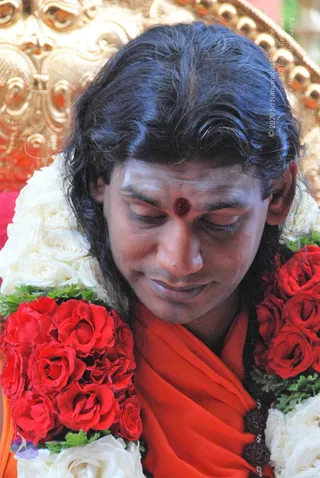 A close photo of a man with long, swept-back dark hair and eyes lowered, wearing an orange-red robe and a dark red bindi on his forehead. He is draped with garlands of red and white roses and green leaves, and dark rudraksha beads show at his chest. Behind him is an ornate gold carved chair or throne. The frame is cropped tightly to his face and shoulders.