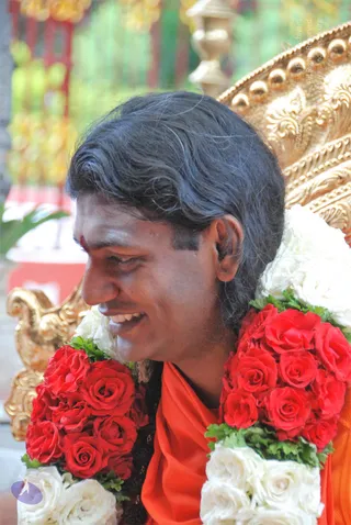 A smiling man with long, swept-back dark hair and a red tilak on his forehead sits in profile before an ornate gold throne. He wears an orange saffron cloth draped over his shoulders and a thick garland of red roses and white flowers with green leaves across his neck. The gilded carved backrest and blurred green and yellow foliage fill the background.