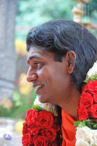 Close side-profile photograph of a smiling man with shoulder-length dark wavy hair and a small red tilak on his forehead. He wears a saffron-orange garment and a heavy garland of red roses interspersed with white roses and green leaves draped over his shoulders. The background is blurred greenery and a pale stone column.