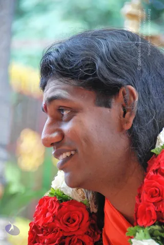 Close profile photograph of a smiling man with shoulder-length dark wavy hair and a red tilak mark on his forehead. He wears a large garland of red roses and white flowers with green leaves over an orange saffron cloth. The background is softly blurred green foliage with yellow blossoms.