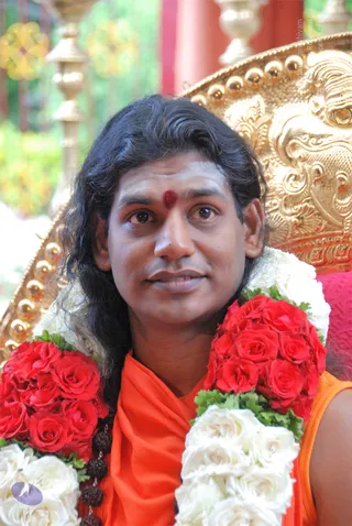 Close-up portrait of a man with long dark wavy hair and a red kumkum dot on his forehead, wearing an orange saffron shawl and draped garlands of red and white roses with rudraksha beads. He sits in front of an ornate gold throne with carved lion-like detailing and a red cushion, behind which stand gold pillars. Swamiji (probable): long swept-back hair, saffron clothing and rudraksha malas indicate the man is.