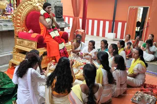 A seated man in saffron-orange robes sits on an ornate gold throne with a red cushioned seat, wearing a large garland of red and white flowers and pressing his palms together in greeting. Around him on the floor, a group of women in white and yellow-bordered saris and a few men in white dhotis sit in a semicircle, several holding brass plates with fruit, sweets, flowers and lit oil lamps. Behind them is an orange-and-white striped wall and a brass lamp stand, indicating a temple-style hall. The man on the throne is a figure presented with honour; the appearance cues of a long-haired figure in saffron clothing and rudraksha are not clearly visible here, so identity is uncertain.