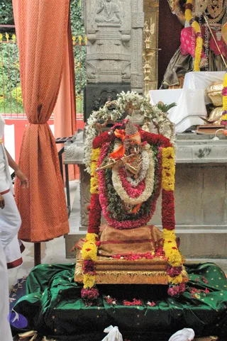 A photograph of a small golden Krishna murti with a flute at its lips, dressed with an orange cloth and a crown, set on a gold-edged base and framed by a dense arch of white jasmine garlands, red and pink flower strands, and yellow marigold garlands hanging down the sides. Flower petals are scattered on a green silk cloth in front. Behind it stand a carved grey stone pillar, a brass oil lamp, and an orange curtain on the left. A figure in white dhoti stands at the left edge. Visible ritual setting suggests puja decoration.