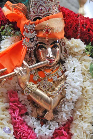 Close-up photograph of a silver-gold Krishna murti adorned for worship. The deity holds a flute to its lips, wears an orange headwrap with a peacock feather and a jewelled crown, and has red and white stone earrings, layered gemstone necklaces and jewelled armlets. Dense garlands of white jasmine and red roses surround the figure, with green leaves at the upper left. No physical people are visible.
